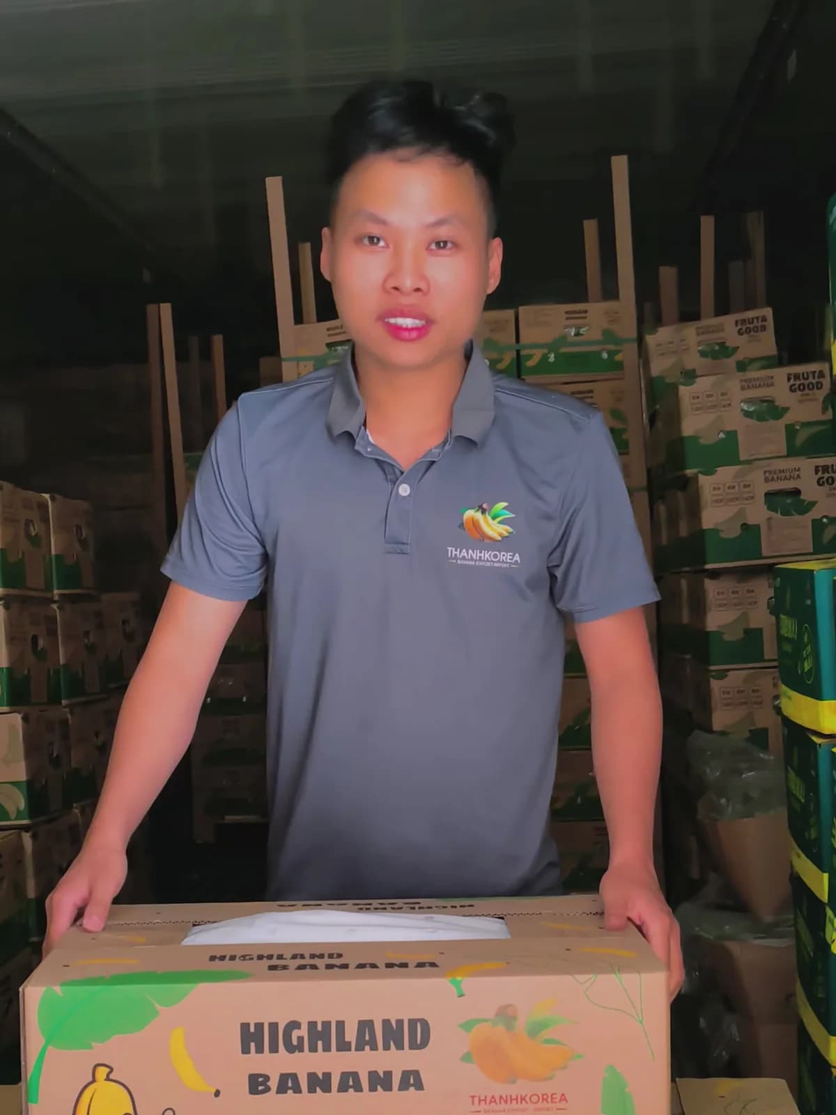 Anh Thành mặc áo THANHKOREA nhìn thẳng ống kính, đứng sau thùng Highland Banana trong kho hàng xuất khẩu
